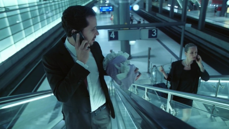 zwei Rolltreppen nach oben, ein Mann übergibt eine Babypuppe an eine Frau